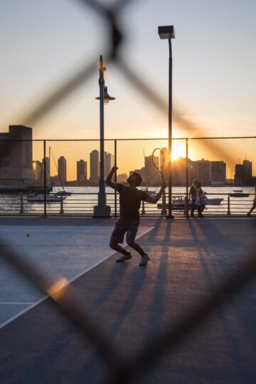 A tennis player begins the serve with the sun setting in the distance