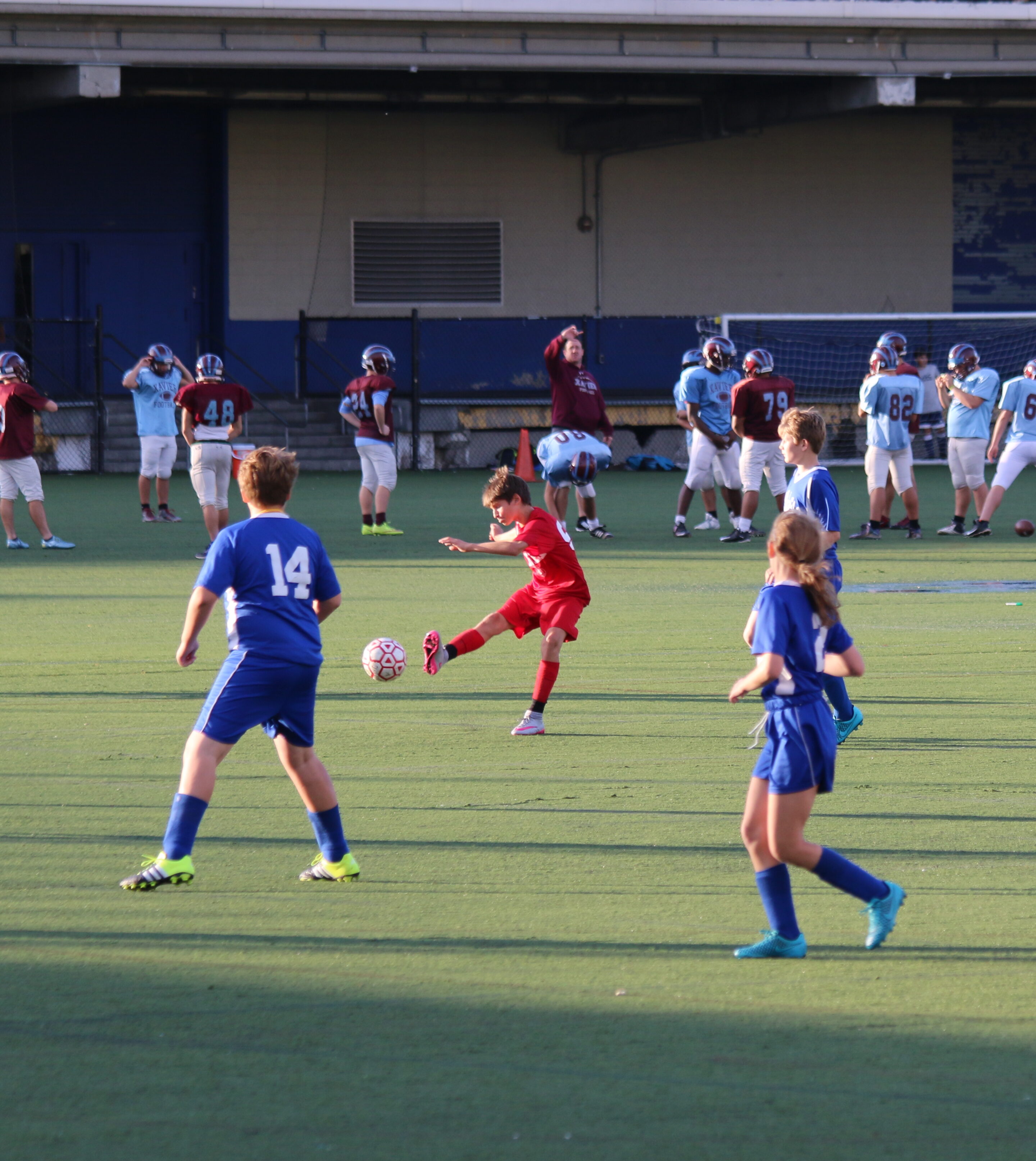 A kid in a red jersey kicks a soccer ball while two kids in blue jerseys defend