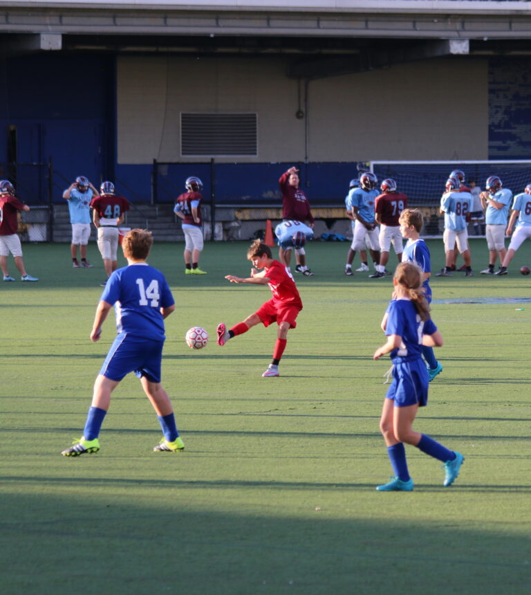 A kid in a red jersey kicks a soccer ball while two kids in blue jerseys defend