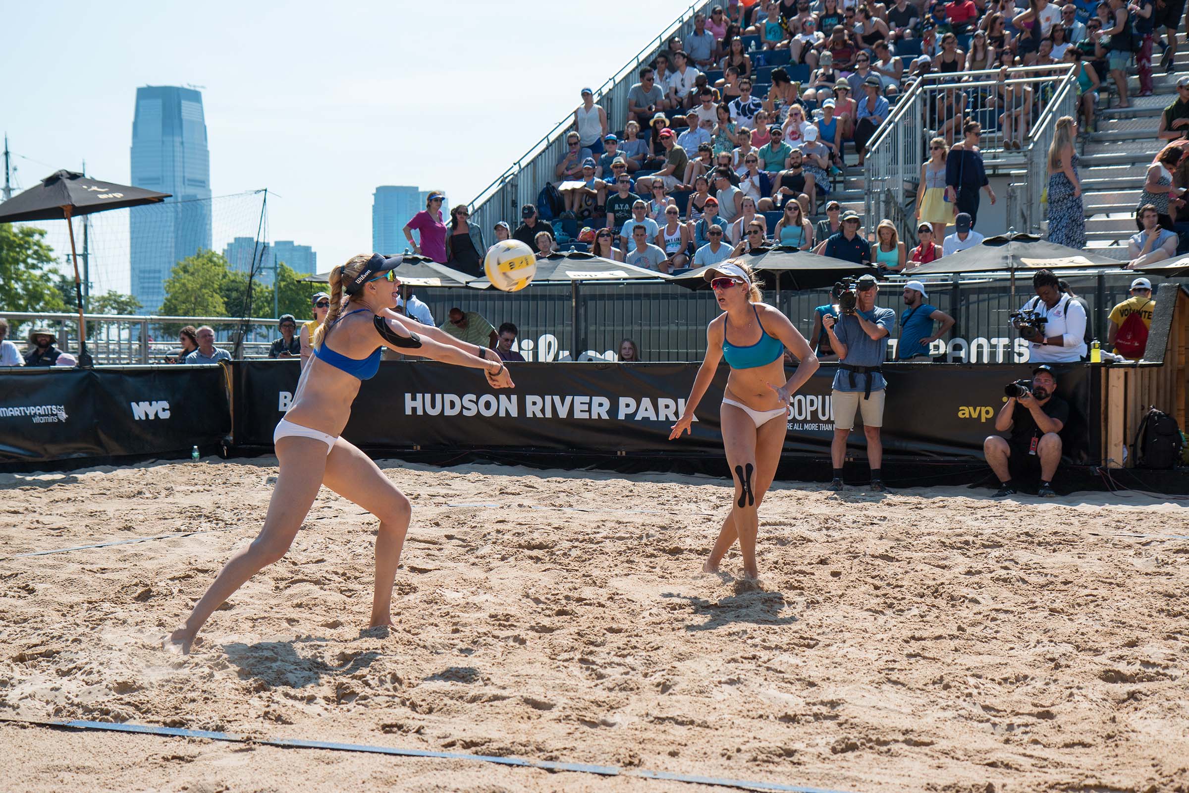 Two beach volleyball players playing in front of an audience in bleachers; one player is performing a dig while the other moves into position for a set.