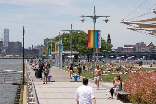 A large gathering of park visitors sit on the green lawn at Pier 45 sunbathing and admiring the views of the Hudson River