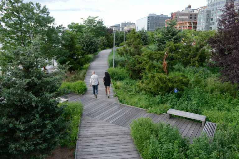 Two Park visitors walk through the Tribeca boardwalk surrounded by lush green trees at Hudson River Park