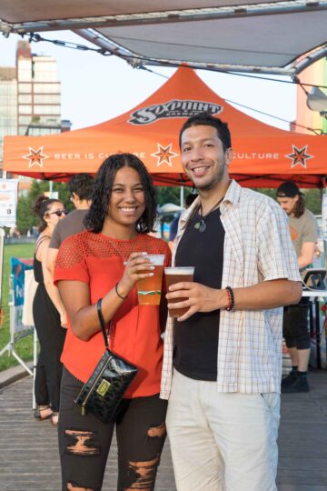 Two Park visitors smiling and holding cups of Sixpoint beer in front of a Sixpoint tent