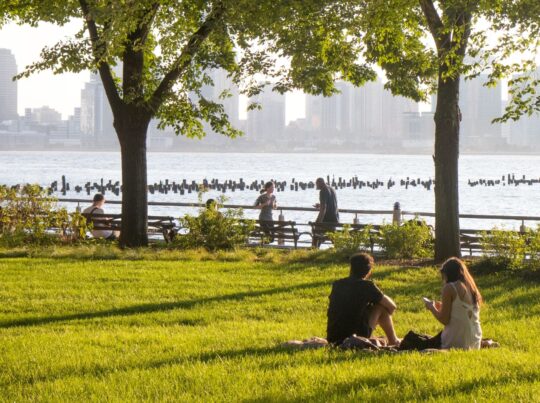 Two Park patrons enjoy a greenwich village picnic on the green Pier 46 lawns with a view of the Hudson River at HRPK