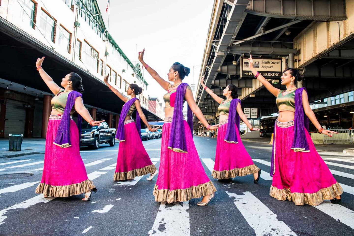 Dancers in pink dresses and purple scarves dance in the street