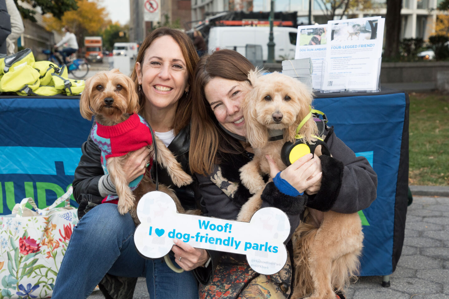 Two women and two dogs pose together with a Hudson River Park Four-Legged Friends sign at HRPK's Barktoberfest