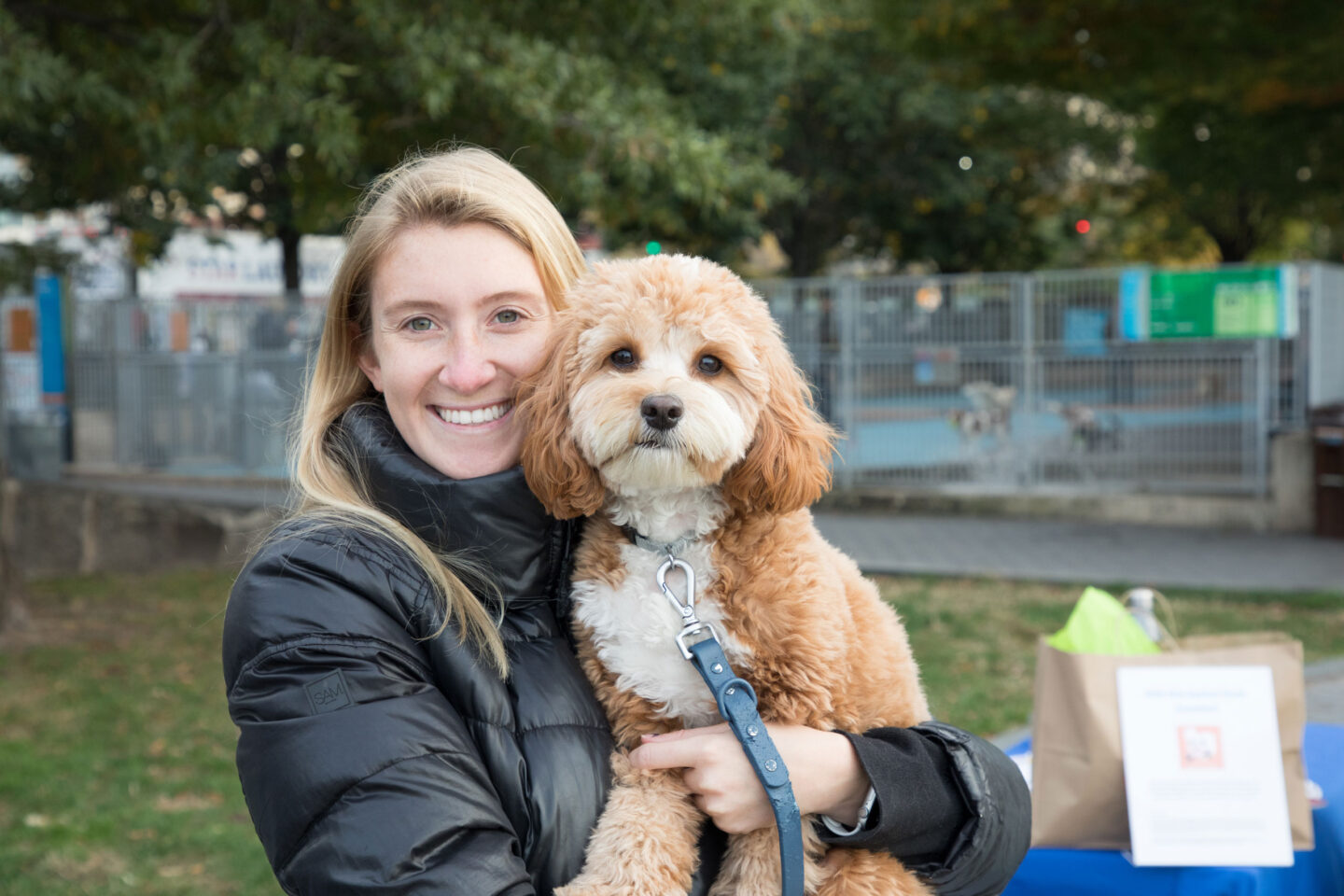 Woman smiles and holds her puppy in front of the Leroy Street Dog Run close at Hudson River Park Friends' Barktoberfest