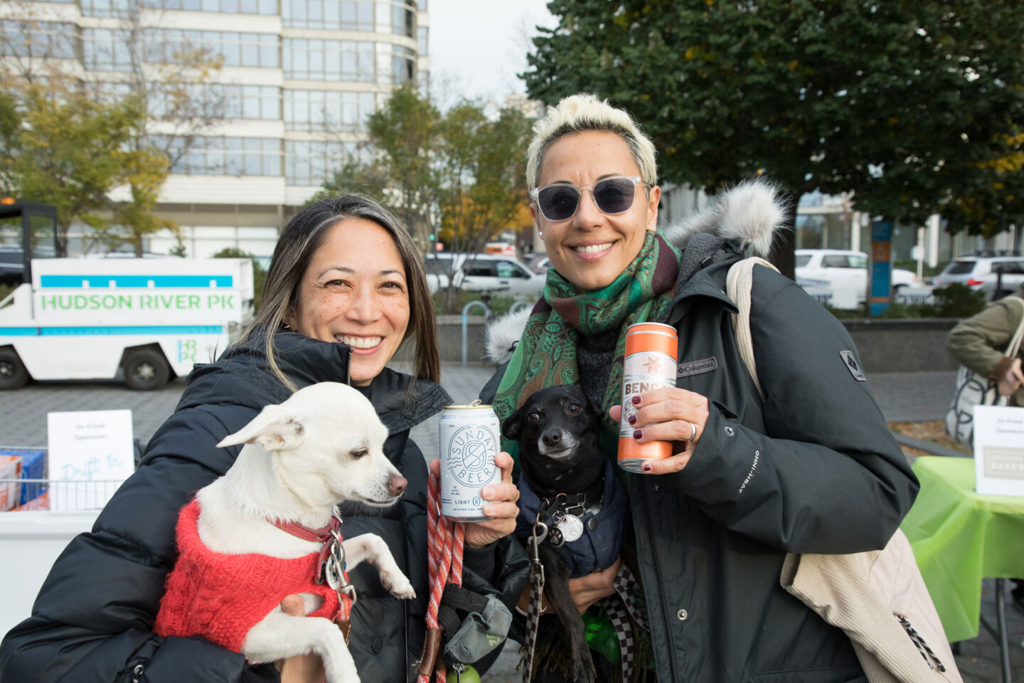 Two smiling women both hold a dog and a sponsored beer in each hand at Hudson River Park Friends' Barktoberfest