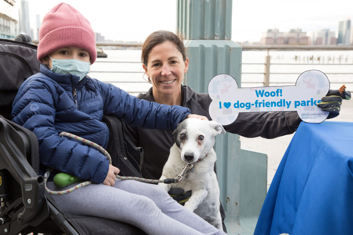 Mom, son and dog hold up a bone-shaped sign promoting Hudson River Park Friends' Four-Legged Friends program against the waterfront at HRPK's Barktoberfest