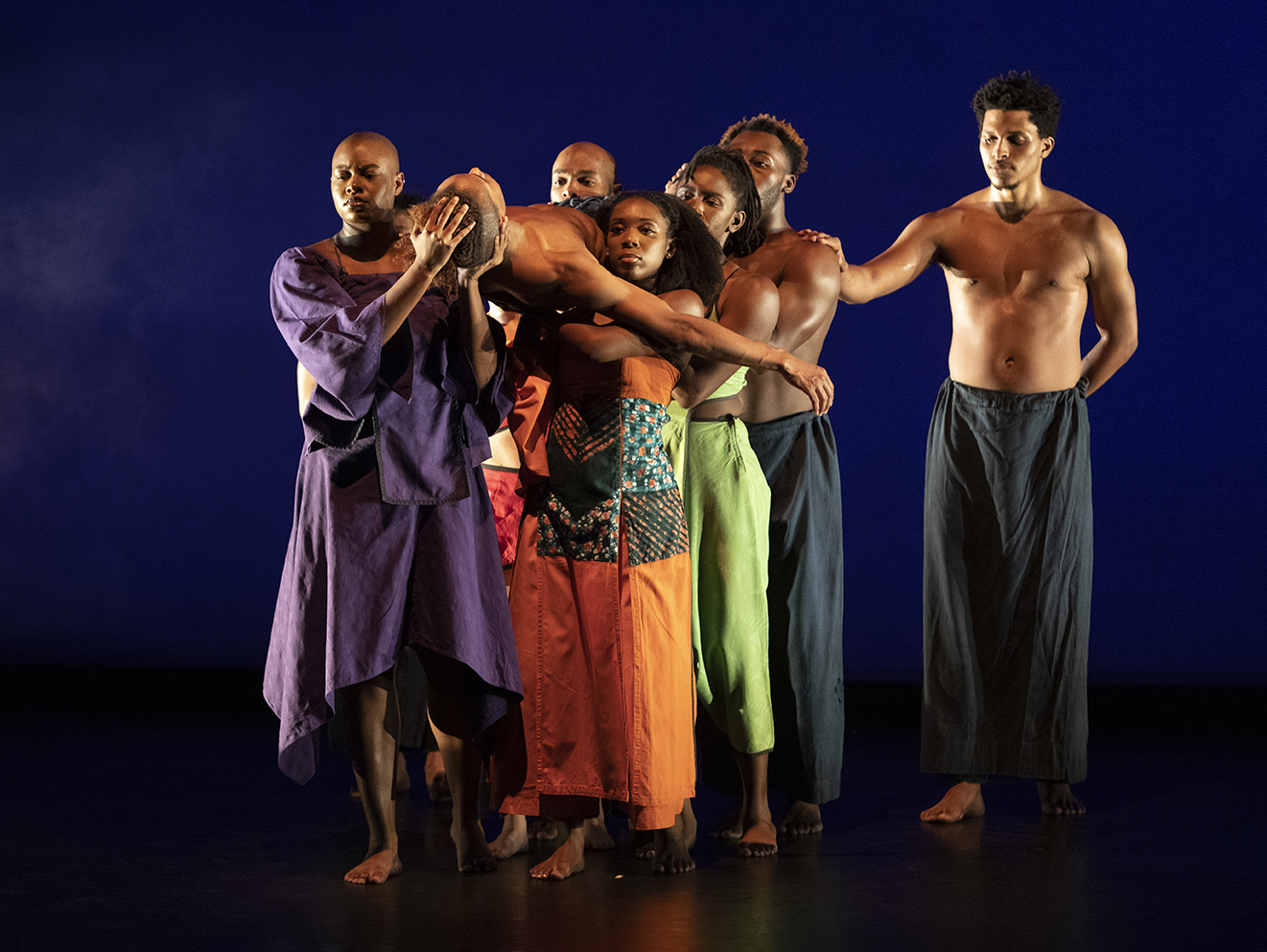 A group of black dancers perform wearing multi colored clothing.