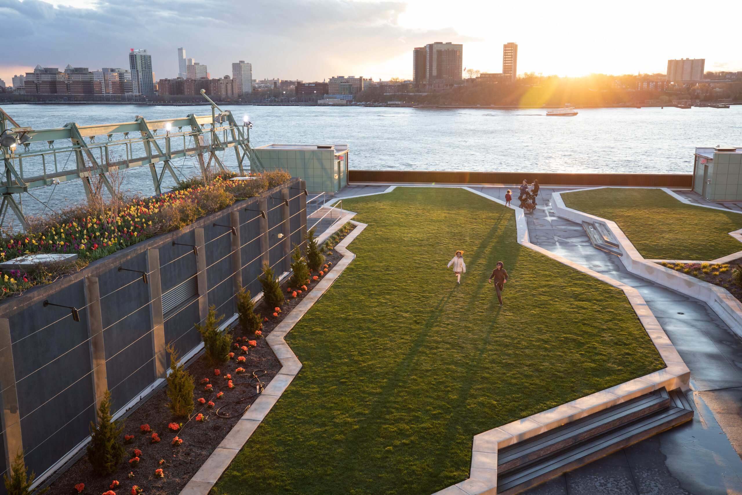 Two children playing on a lawn on the Pier 57 rooftop, while a group walks along a walkway between lawns.