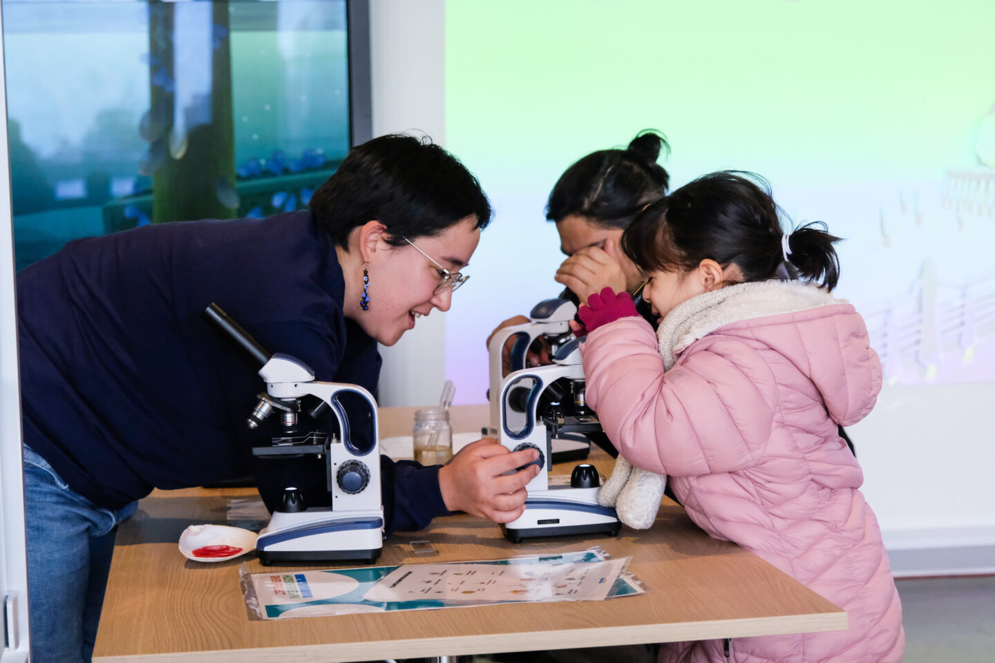 A child and their parent look through microscopes while an HRPK River Project staffer explains to them what they're seeing