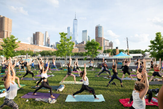 Outdoor yoga class in Hudson River Park with participants on mats, arms raised, and the Lower Manhattan skyline in the background.