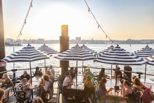 Groups of people sitting at tables underneath umbrellas on the Hudson River Park waterfront