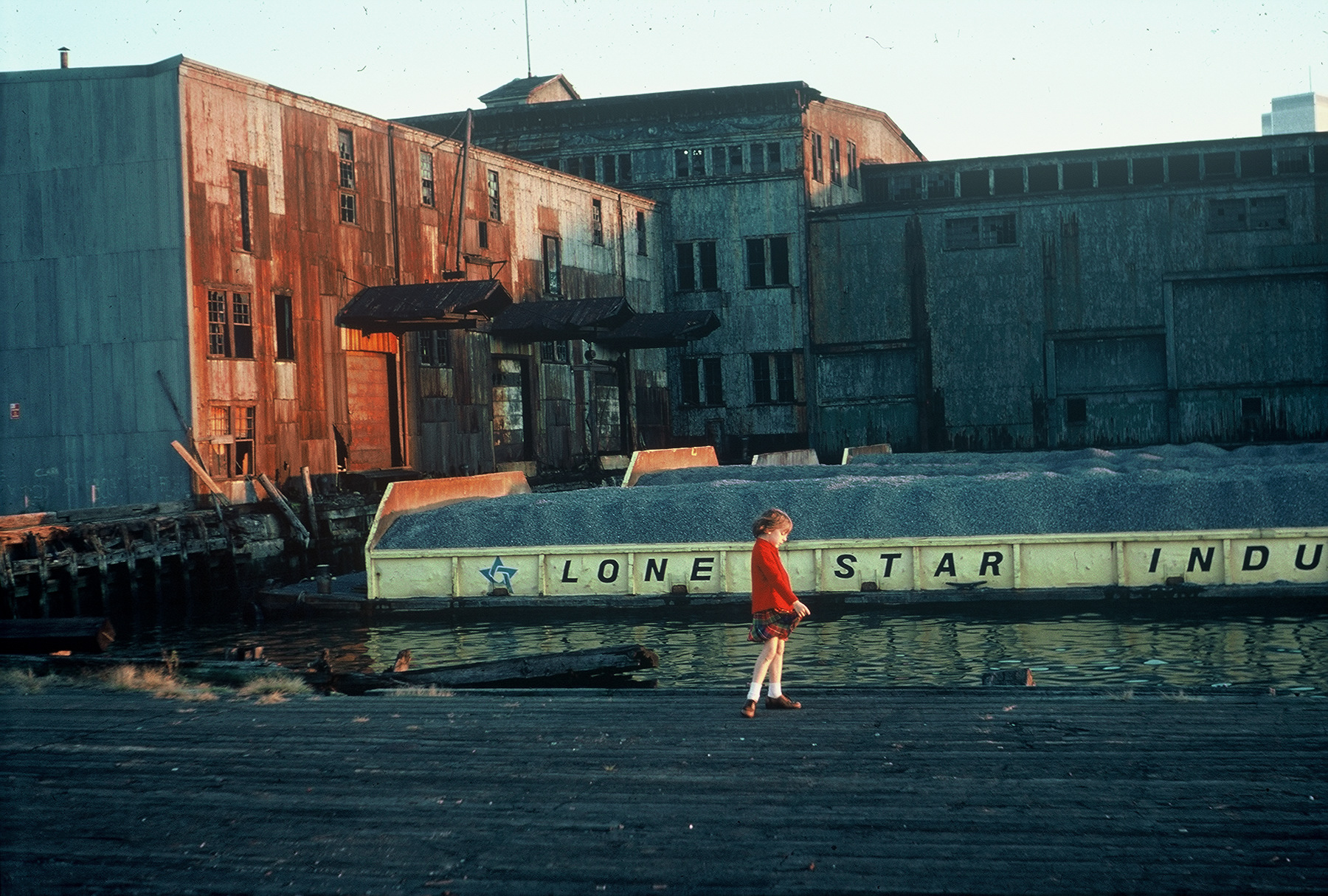 A young girl in a red jacket and plaid skirt standing on a pier with rusting industrial buildings in the background.