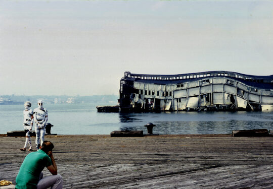 Two people wrapped in bandages like mummies walk on a Hudson River pier. A collapsing pier is visible in the background.