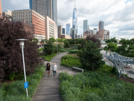 Two people walking on the Tribeca Boardwalk in Hudson River Park. One World Trade Center is visible in the background.