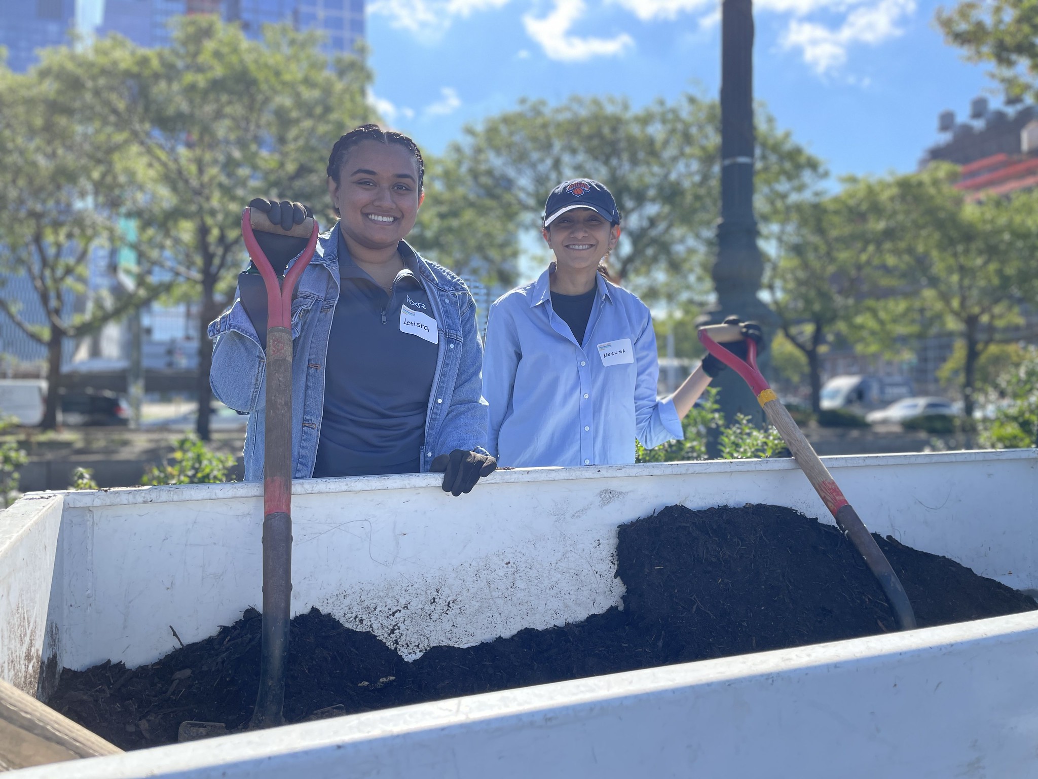 Two volunteers holding shovels pose for a photo behind a mulch container