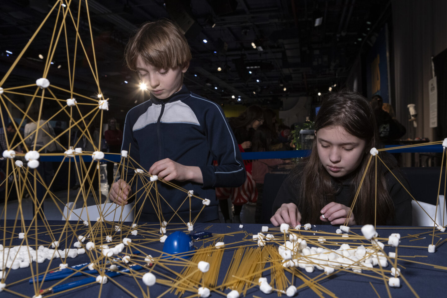 Two students build structural models out of spaghetti and marshmallows at a table inside the Intrepid Museum
