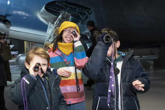 Three children look through small telescopes at an Intrepid Museum exhibit