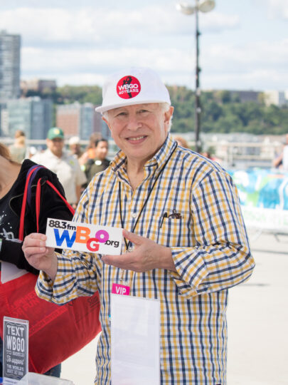 A person holding a WBGO 88.3 FM sticker ad wearing a "WBGO 40 Years" hat at a Blues BBQ Festival table
