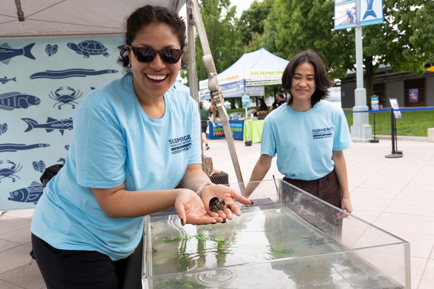 Two HRPK River Project staffers stand at an aquarium touch tank booth at the SUBMERGE Marine Science Festival on Pier 84. One staffer is holding a small crab in their hands.