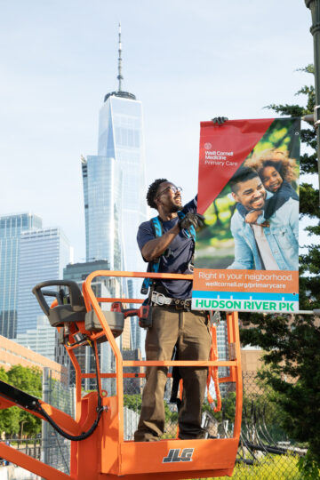 A HRPK staffer hangs a lightpole banner advertising Weill Cornell Medicine Primary Care along the Hudson River Greenway; One World Trade Center is visible in the background.
