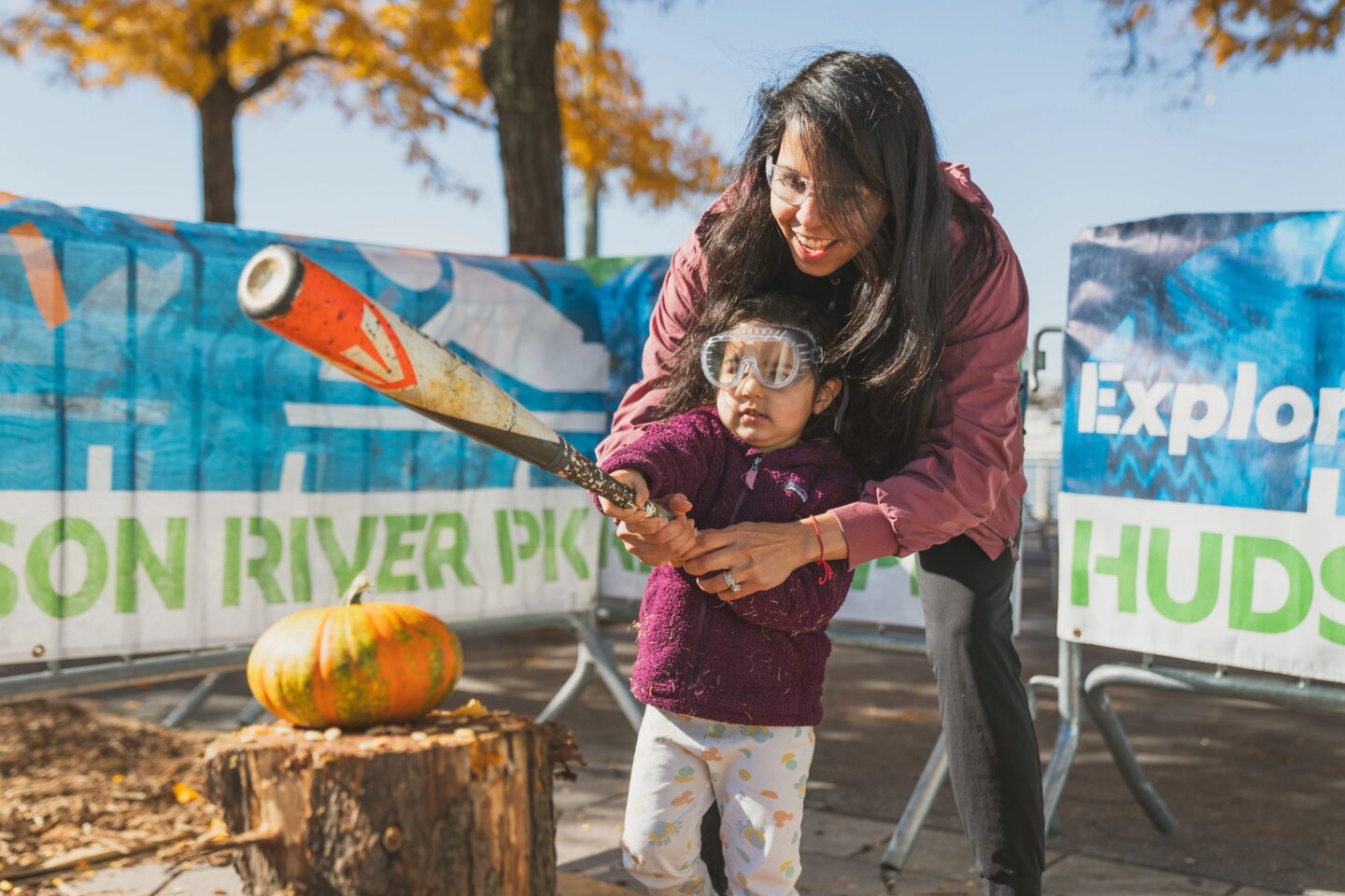 A parent and child swinging a baseball bat down at a pumpkin on a stump, at the Pumpkin Smash event at Pier 84