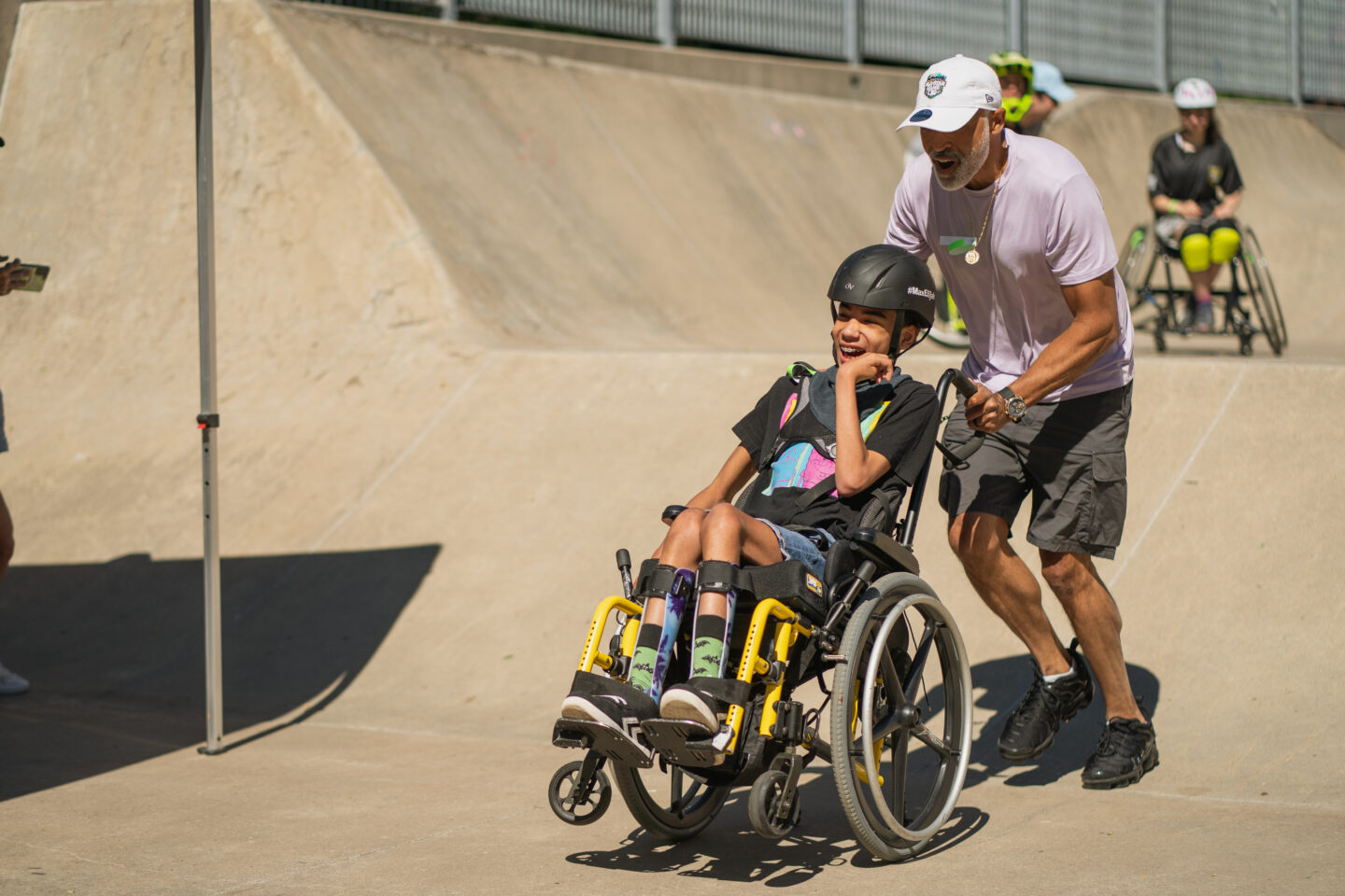 An adult pushes a child in a wheelchair down a ramp at the Pier 62 Skatepark