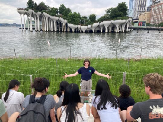 An HRPK River Project staffer teaches a class of students in front of the Gansevoort Peninsula Salt Marsh, with Little Island visible in the background