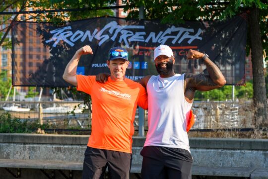 Dr. Jordan Metzl and an IronStrength class instructor do a flexing pose in front of an IronStrength banner on Pier 25