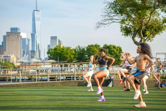 A fitness instructor leads a Healthy on the Hudson class in a "running in place" exercise on the Pier 46 turf field. One World Trade Center is visible in the distance.