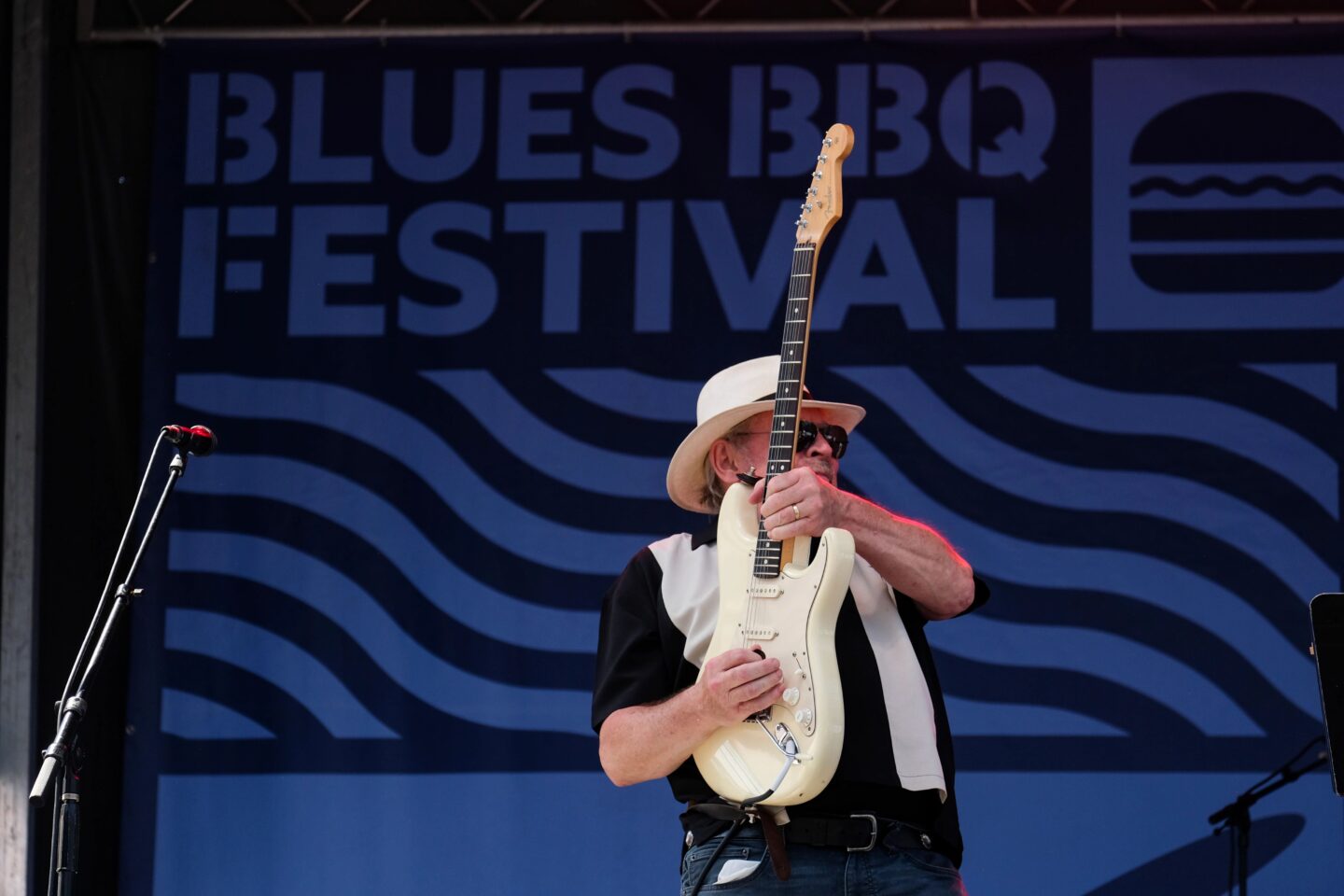 A guitarist holds his guitar vertically while playing in front of a backdrop that reads "Blues BBQ Festival" at a free music festival in NYC
