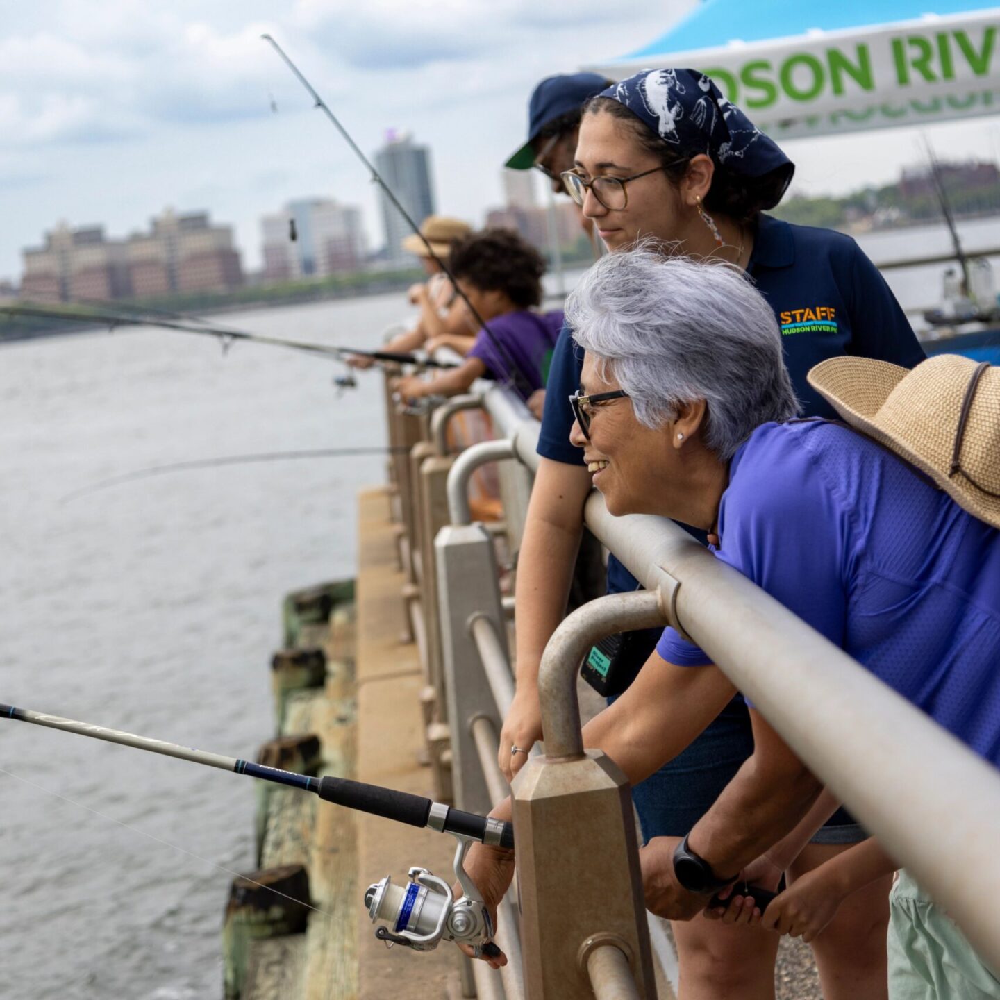 Children and adults hold fishing rods off the edge of a Hudson River Park pier during a Big City Fishing event