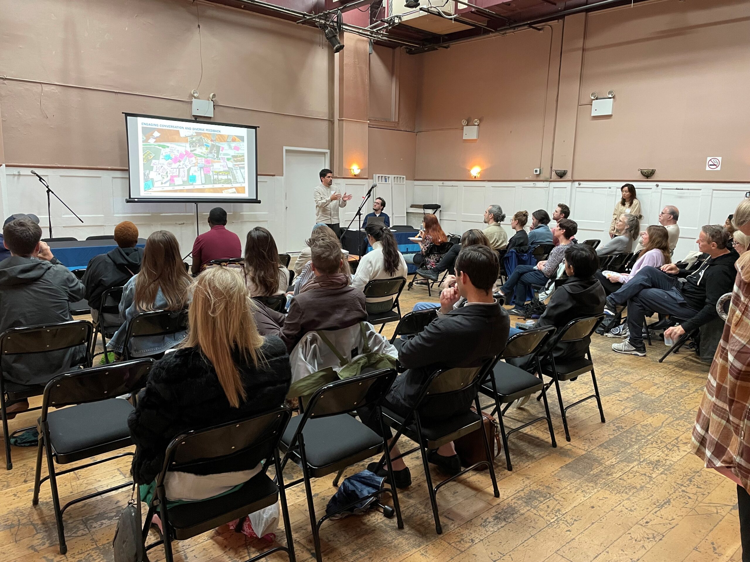 A presenter from the engineering firm Marvel guides a seated audience through a slide deck presentation at a public meeting