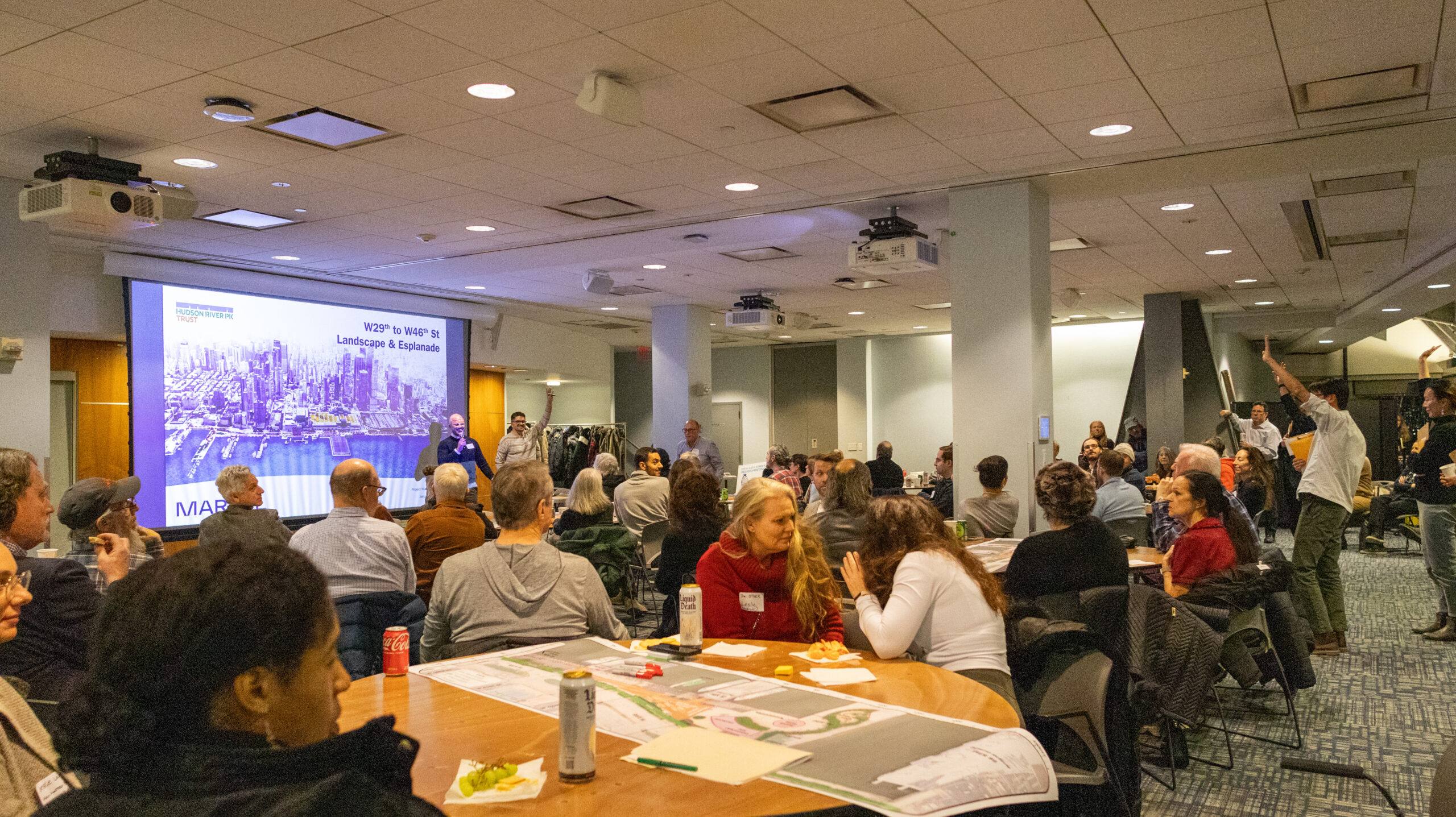 Tables filled with members of the local community watch a presentation on a projector screen during A presenter speaks in front of a projector screen during a W 29–W 44 Esplanade & Landscape Engagement Meeting at the Intrepid Museum