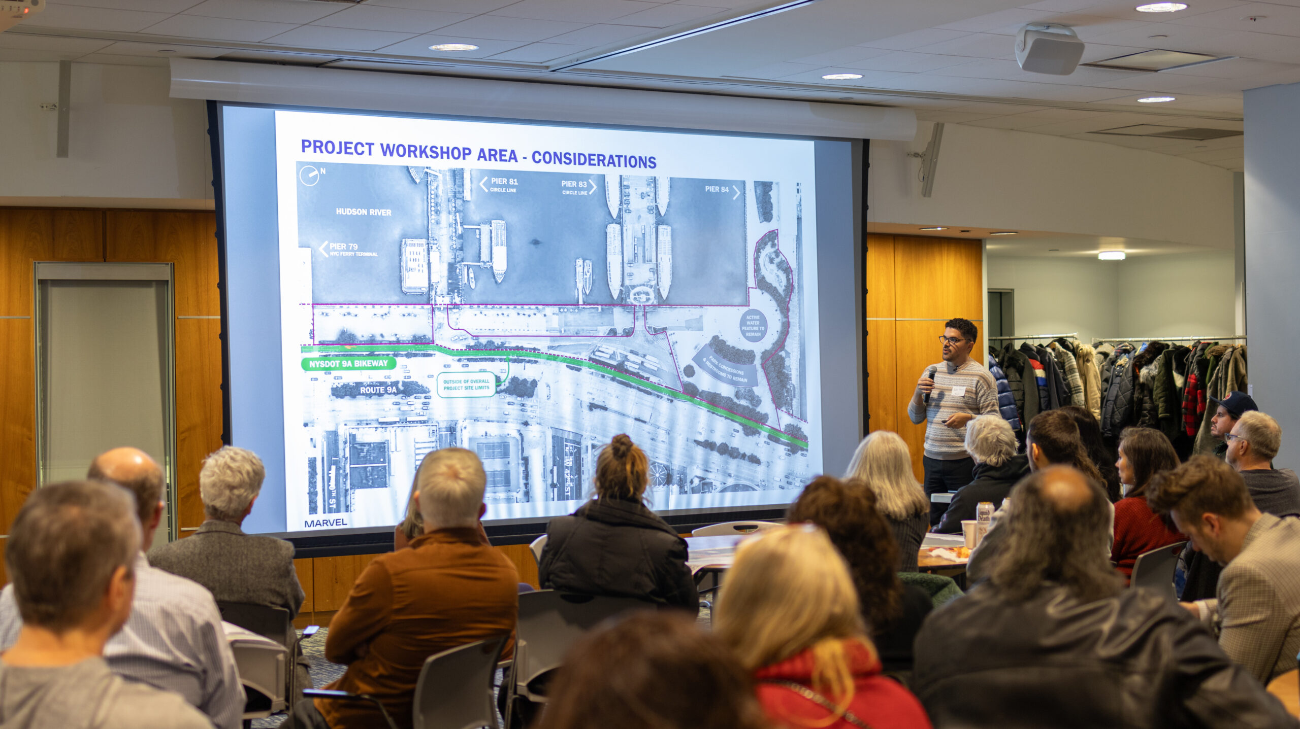 A presenter speaks in front of a projector screen during a W 29–W 44 Esplanade & Landscape Engagement Meeting at the Intrepid Museum