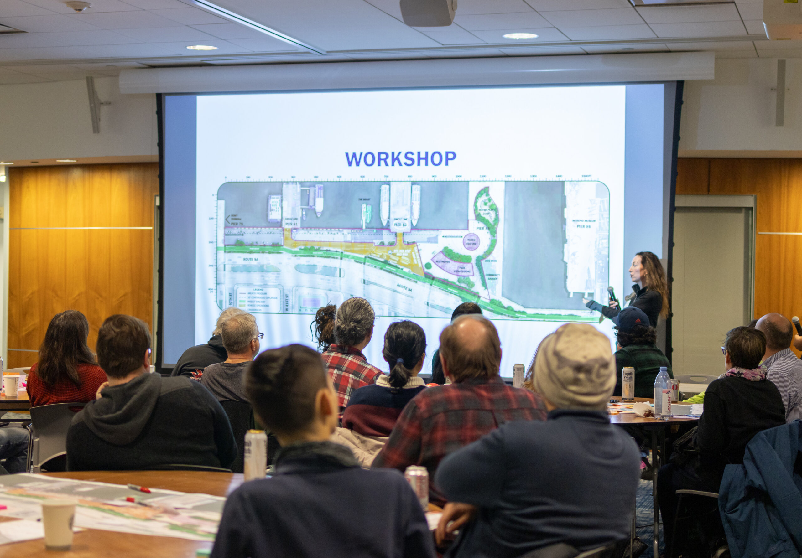 A presenter speaks in front of a projector screen during a W 29–W 44 Esplanade & Landscape Engagement Meeting at the Intrepid Museum