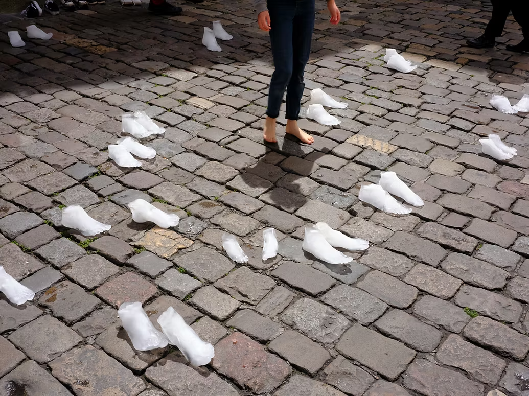 A person standing barefoot on cobblestones, surrounded by molds of feet frozen in ice