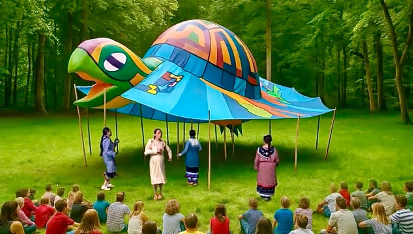 Performers dance on a lawn underneath a large, colorful inflatable turtle