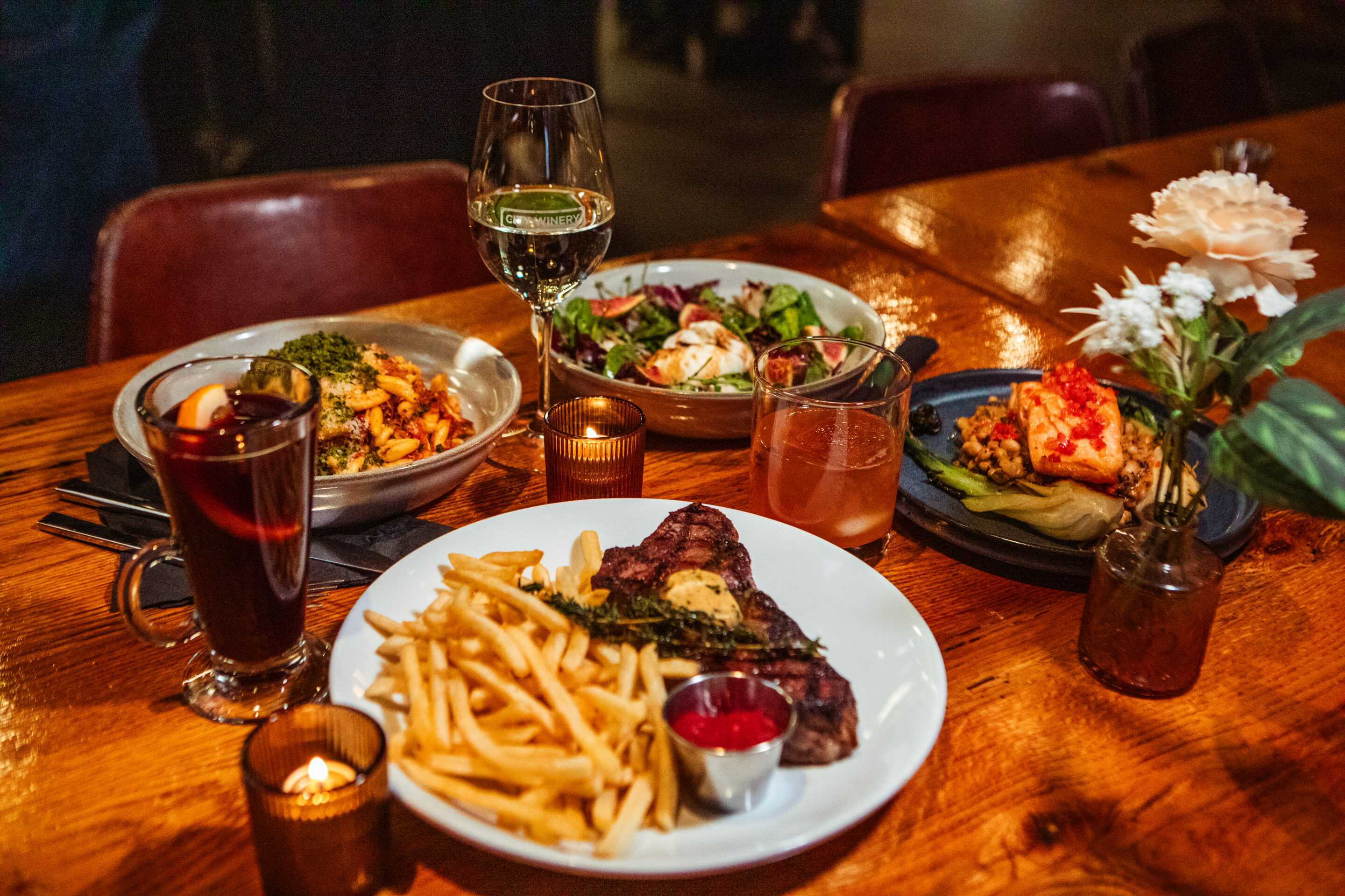 A City Winery table setting with four dinner plates—a steak dish with fries, a pasta dish, a salad, and a seafood dish—accompanied by two cocktail glasses and a glass of wine