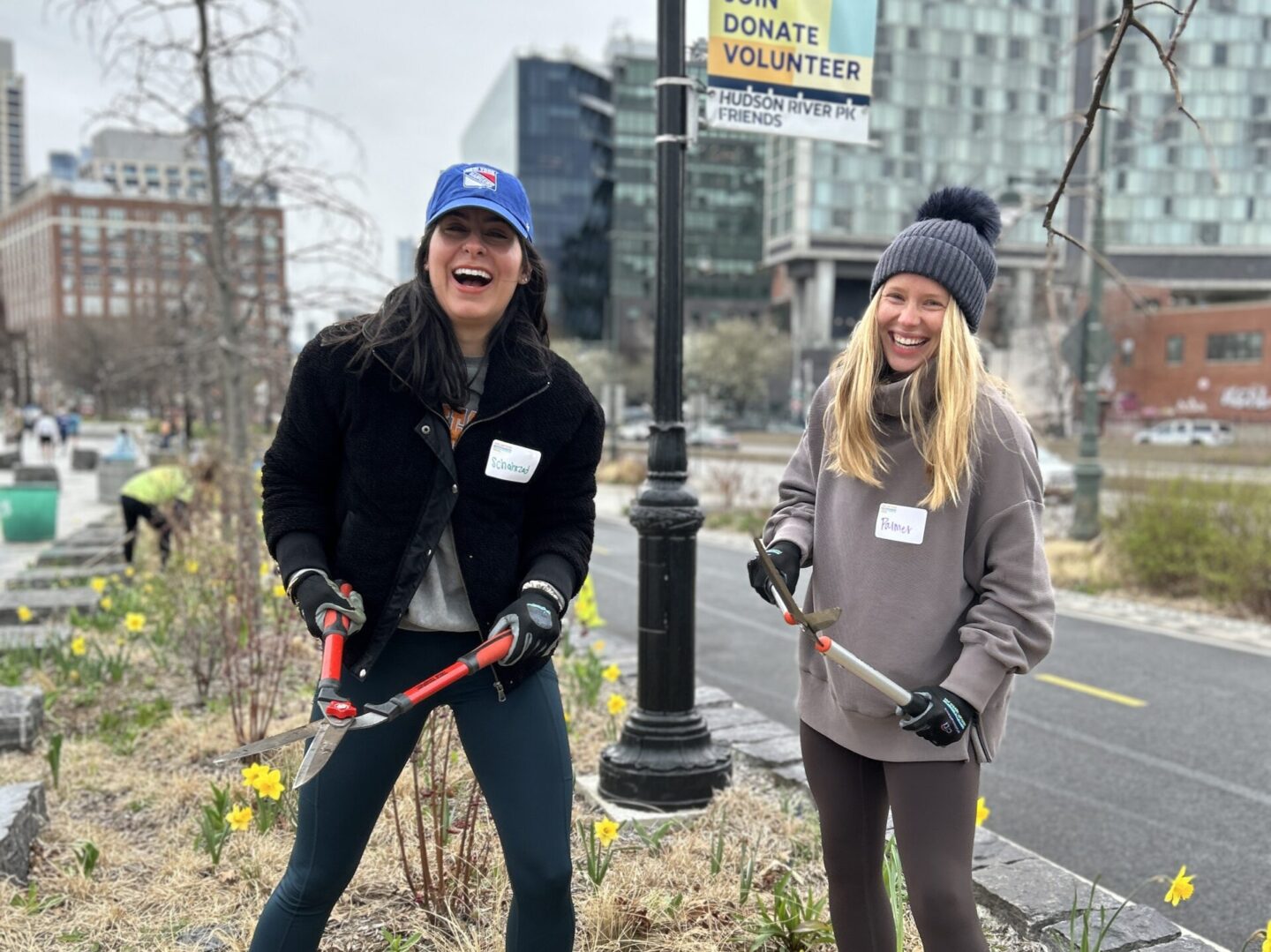 Two women holding gardening tools in on a cloudy day on top of some grasses in Hudson River Park