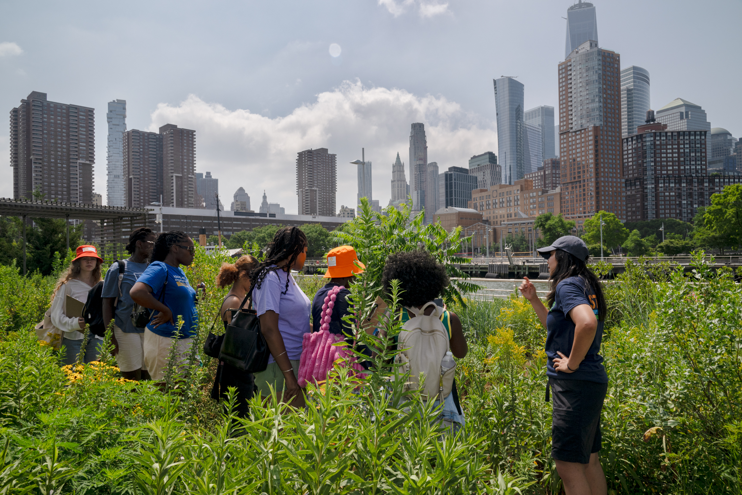 Group tour in a Hudson River Park garden with a staff member speaking to visitors, surrounded by native plants and Lower Manhattan buildings.