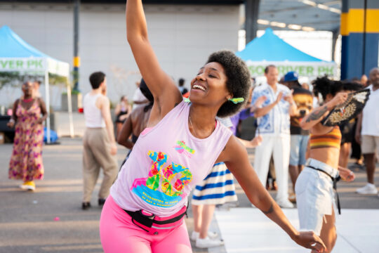Smiling participant dancing with one arm raised during an outdoor Hudson River Park event.
