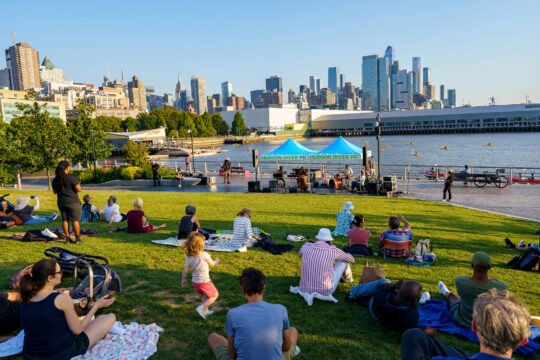 Audience watching a live blues performance on the Hudson River Park lawn, with the Hudson River and Manhattan skyline in the background.