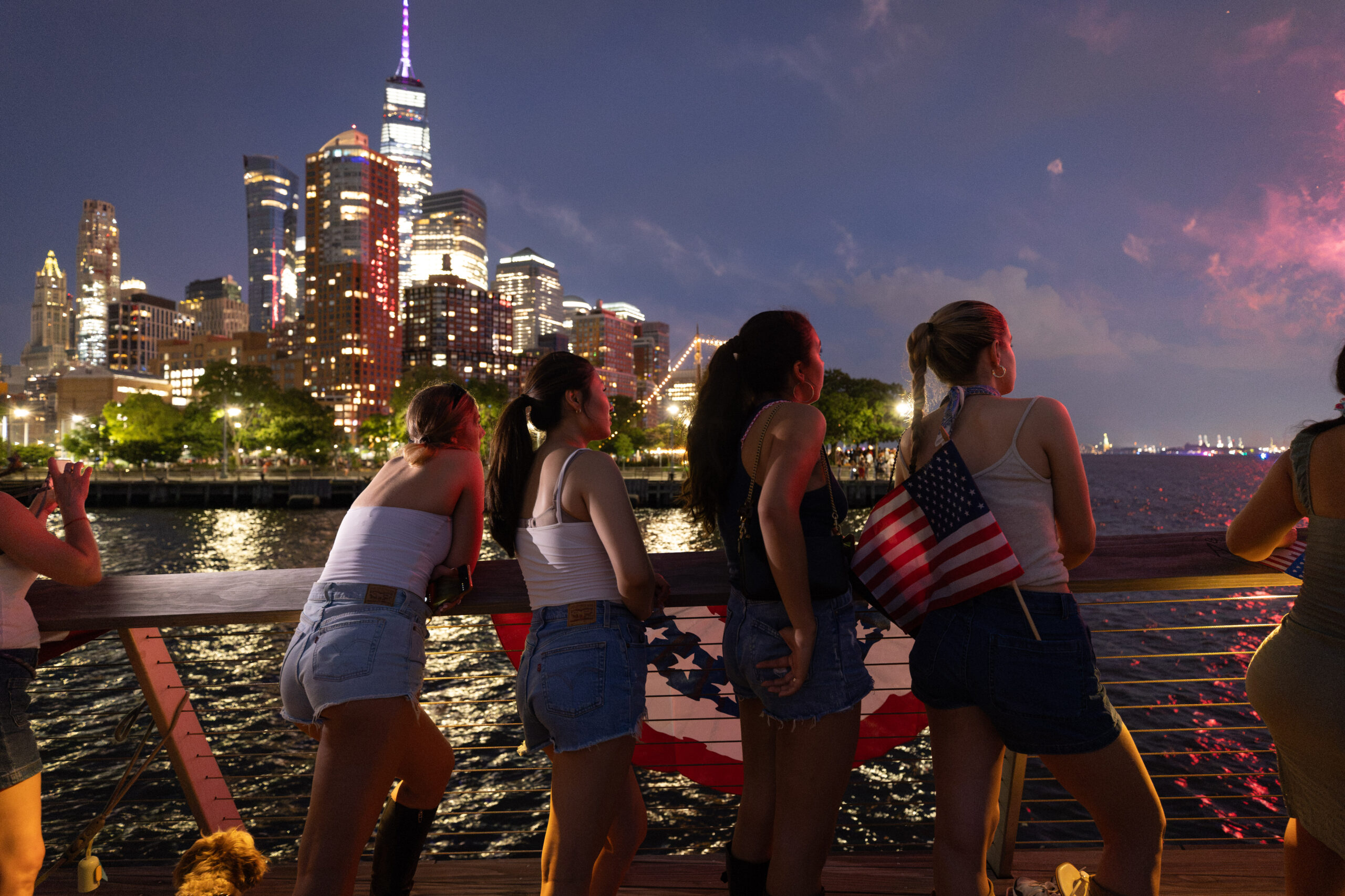 young women stand at the edge of Pier 26 at night for fireworks; Lower Manhattan lit up in background