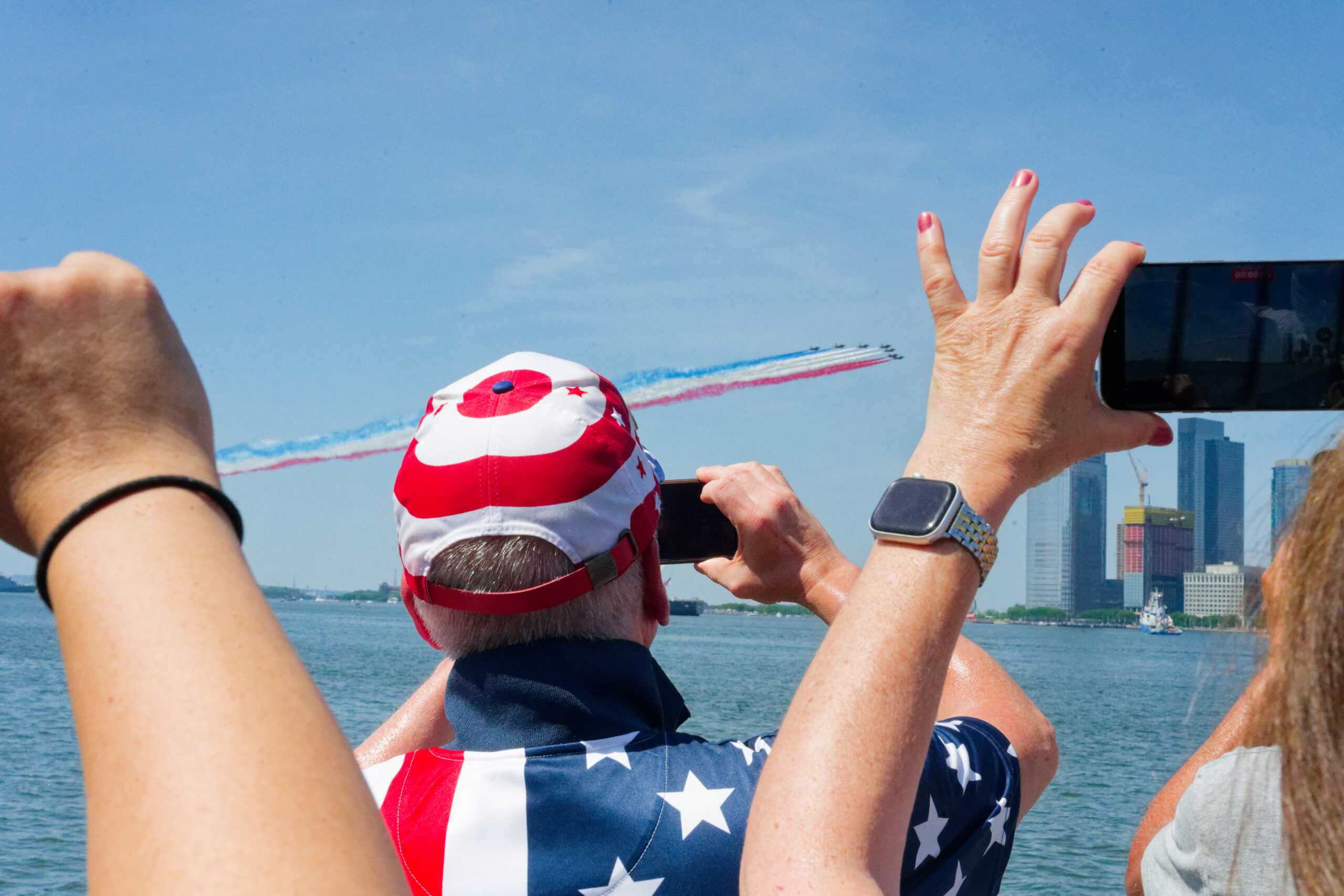 People hold phones up to capture view of planes trailing red white and blue overhead