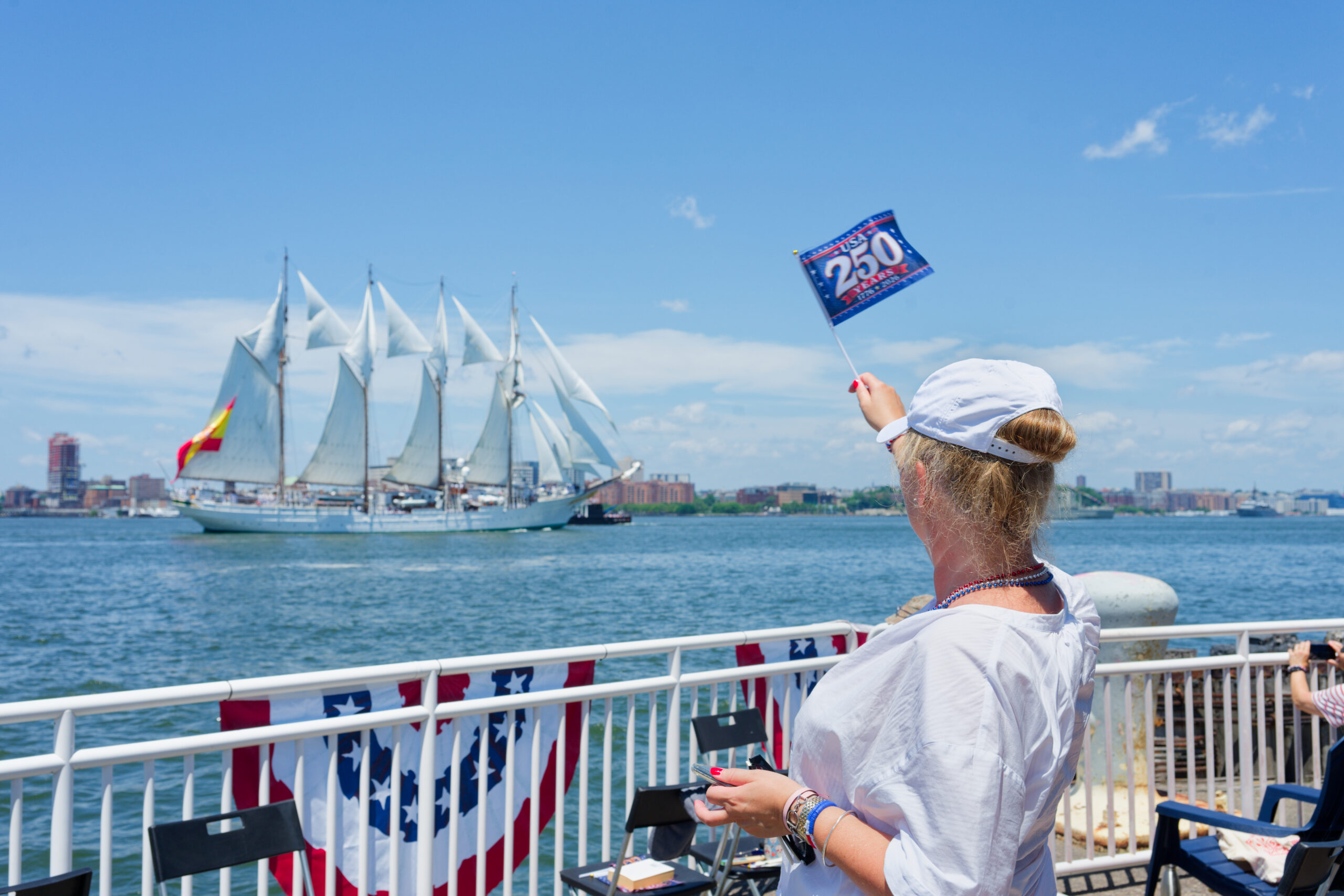 Woman stands by railings by the water in white hat waves an America's 250th flag as a tall ship sails the Hudson River