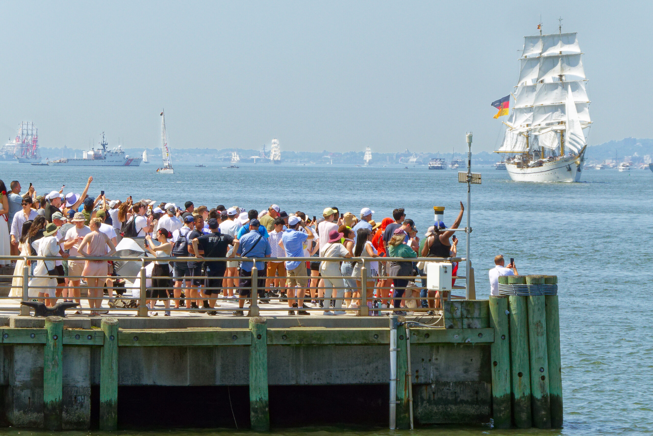 Crowd of people at edge of pier with tall ship on the river in the background