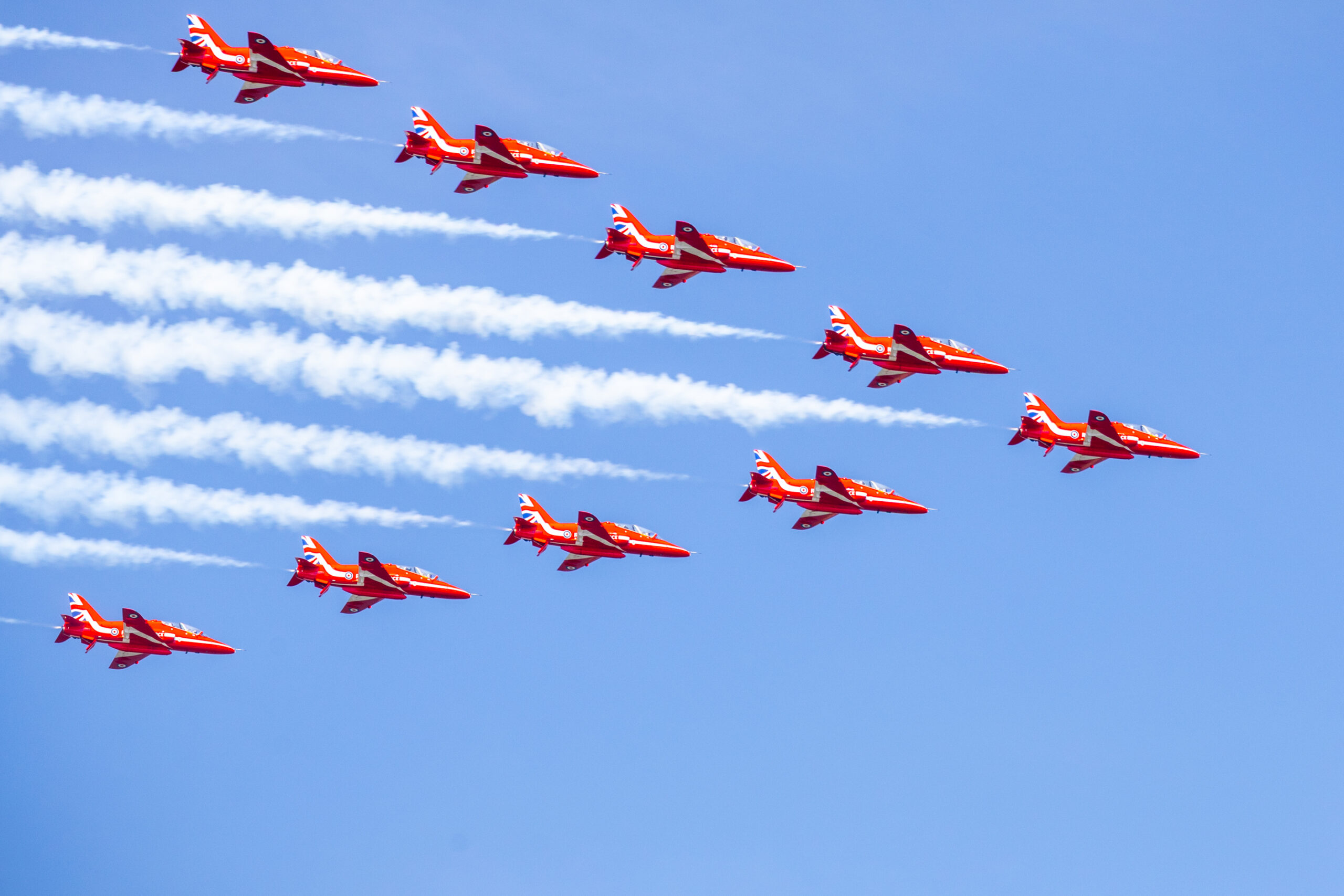 Red planes in v formation fly overhead against blue skies leaving white trails